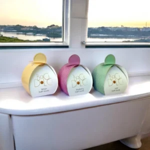 Three pastel-colored bath bomb boxes with a curved, dome-like shape and a quilted texture. The packaging is labeled "Baci Dolce" and features gold-foil typography, placed on a bathtub with a scenic outdoor view.